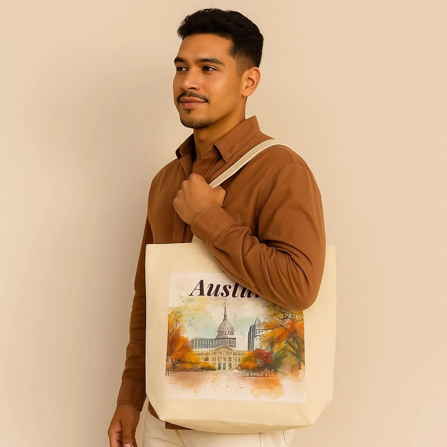Stylish young man with medium brown skin wearing a beige sweatshirt, holding a beige organic cotton tote bag with Austin city illustration on the shoulder, neutral background.