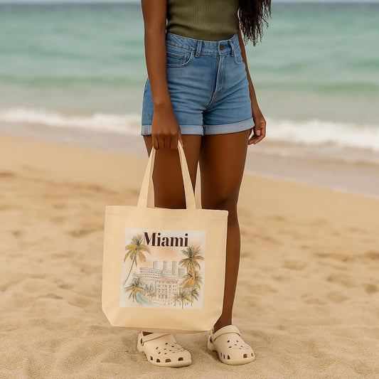 A woman stands on the beach holding a Miami tote bag by its handles. The ocean and sand create a relaxed backdrop. She's wearing denim shorts and Crocs. The bag features a tropical watercolor design of Miami architecture and palm trees.